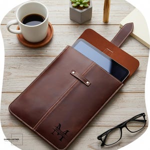May include: A brown leather tablet case with a personalized monogram. The case is open, revealing a tablet inside. The case has a stitched design and a strap closure. A coffee cup, coaster, and glasses are on the wooden surface.