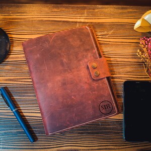 K&ouml;nnte beinhalten: Ein braunes Lederjournal mit einem Klappverschluss und einem Kreis, der mit den Initialen "MR" gepr&auml;gt ist. Das Journal liegt auf einer Holzoberfl&auml;che mit einem schwarzen Stift, einem goldenen Dekorationsobjekt und einem Strau&szlig; getrockneter Blumen.