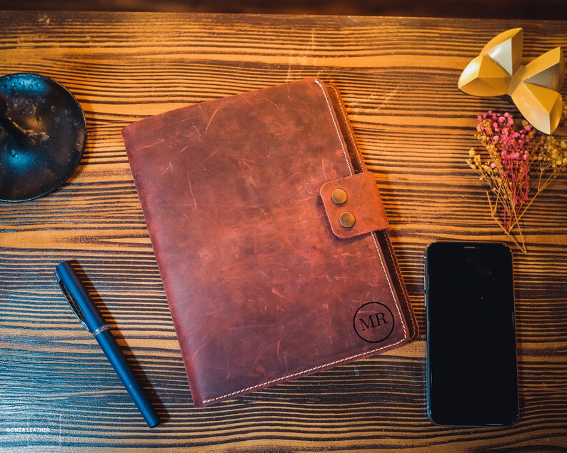K&ouml;nnte beinhalten: Ein braunes Lederjournal mit einem Klappverschluss und einem Kreis, der mit den Initialen "MR" gepr&auml;gt ist. Das Journal liegt auf einer Holzoberfl&auml;che mit einem schwarzen Stift, einem goldenen Dekorationsobjekt und einem Strau&szlig; getrockneter Blumen.