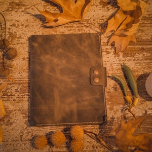 K&ouml;nnte beinhalten: Ein braunes Ledernotizbuch mit rustikalem Aussehen und zwei Messingnieten auf dem vorderen Umschlag. Das Notizbuch liegt auf einer Holzoberfl&auml;che mit Herbstbl&auml;ttern und anderen nat&uuml;rlichen Elementen, die um es herum verstreut sind.