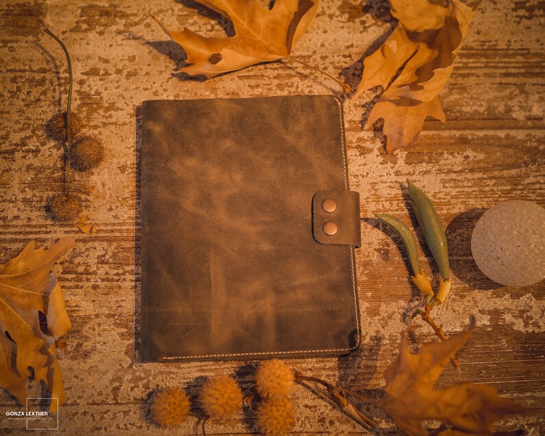 K&ouml;nnte beinhalten: Ein braunes Ledernotizbuch mit rustikalem Aussehen und zwei Messingnieten auf dem vorderen Umschlag. Das Notizbuch liegt auf einer Holzoberfl&auml;che mit Herbstbl&auml;ttern und anderen nat&uuml;rlichen Elementen, die um es herum verstreut sind.