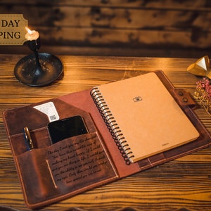 May include: A brown leather journal with a spiral bound notebook inside. The journal has a pocket on the front cover with a phone and pen inside. The journal is open to a blank page with a small camera icon printed on it. The journal is on a wooden table with a lit candle and a gold decorative object in the background. The text "The day I met you my life changed. You have this incredible way of making my heart happy. If I could give you one thing in life I would give you the ability to see yourself through my eyes. Only then would you realize how special you are to me." is engraved on the front cover.