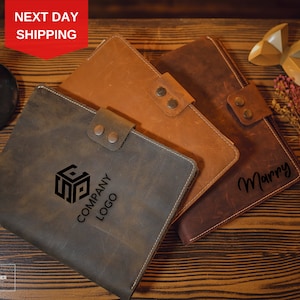 May include: Three leather-bound journals with different designs. One journal has a company logo, another has the word "Marry" in cursive, and the third has a simple design. The journals are on a wooden surface.