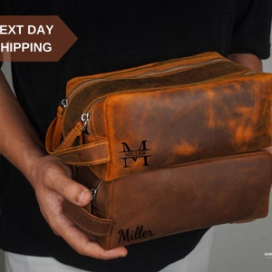 May include: Two brown leather travel bags with a zipper closure. The top bag has a monogrammed "M" with "Miller" underneath. The bottom bag has the name "Miller" engraved on the front.