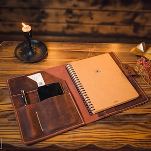 K&ouml;nnte beinhalten: Ein braunes Leder-Notizbuchcover mit einem spiralgebundenen Notizbuch im Inneren. Das Cover hat eine Tasche f&uuml;r ein Telefon und einen Stift. Das Notizbuch ist geschlossen und hat ein Kamerasymbol auf der Vorderseite.