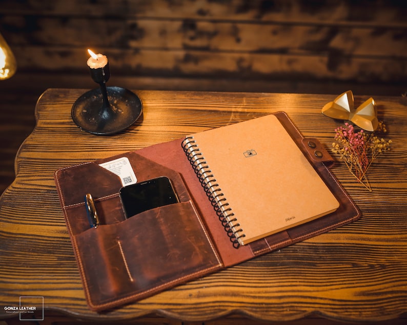 K&ouml;nnte beinhalten: Ein braunes Leder-Notizbuchcover mit einem spiralgebundenen Notizbuch im Inneren. Das Cover hat eine Tasche f&uuml;r ein Telefon und einen Stift. Das Notizbuch ist geschlossen und hat ein Kamerasymbol auf der Vorderseite.