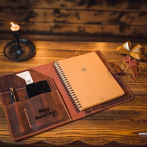 K&ouml;nnte beinhalten: Ein braunes Lederjournal mit einem spiralgebundenen Notizbuch im Inneren. Das Journal hat eine Tasche auf der Vorderseite mit einem Stift und einem Telefon darin. Das Journal ist ge&ouml;ffnet und die Innenseite des vorderen Deckels tr&auml;gt die Worte "Happy Anniversary!" in goldener Pr&auml;gung.