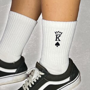 Könnte beinhalten: Weiße Socken mit einem schwarzen gestickten Design eines Spielkarten-Kämpfers von Pik mit einer Krone oben.