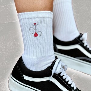Könnte beinhalten: Weiße Socken mit einem roten und grauen gestickten Wasserpfeifen-Design. Die Socken werden mit schwarzen und weißen Sneakern getragen.