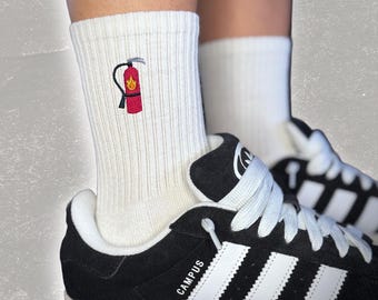 May include: White crew socks with an embroidered red fire extinguisher with a yellow flame. The socks are being worn with black and white sneakers that say "CAMPUS" on the side.