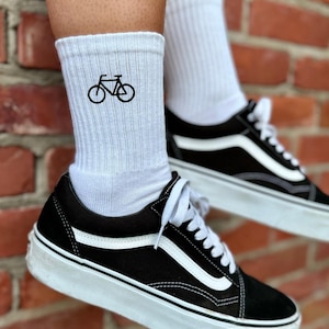 Könnte beinhalten: Weiße Sportsocken mit einem schwarzen Fahrradmotiv, getragen mit schwarz-weißen Turnschuhen. Die Turnschuhe haben ein Obermaterial aus schwarzem Wildleder, einen weißen Seitenstreifen und eine weiße Sohle. Die Socken sind gerippt und reichen bis zur Mitte der Wade.