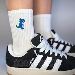 Könnte beinhalten: Weiße Sportsocken mit einem blauen und grünen Dinosaurier-Design. Die Socken werden mit schwarz-weißen Turnschuhen getragen. Die Turnschuhe haben weiße Schnürsenkel und drei weiße Streifen. Das Wort "CAMPUS" ist auf der Seite des Schuhs zu sehen.