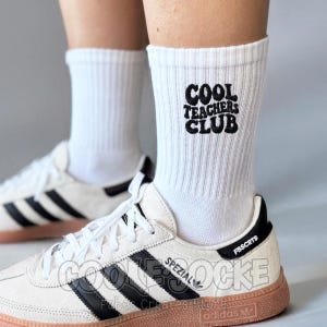 Könnte beinhalten: Ein Paar weiße, gerippte Crew-Socken mit dem schwarzen, gestickten Text COOL TEACHERS CLUB an der Außenseite. Die Socken werden mit weiß-schwarzen Turnschuhen kombiniert, die eine gummifarbene Sohle und weiße Schnürsenkel aufweisen.