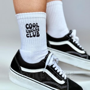 Könnte beinhalten: Weiße Sportsocken mit dem Text "COOL UNCLES CLUB" in Schwarz. Die Socken werden mit schwarz-weißen Turnschuhen getragen.