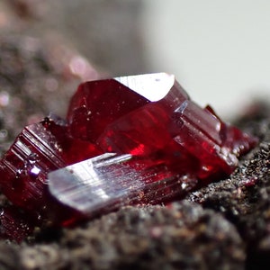 Może przedstawiać: Zbliżenie czerwonego kryształu mineralnego na ciemnoszarym kamieniu. Kryształ ma ostry, fasetowany kształt i błyszczące wykończenie.