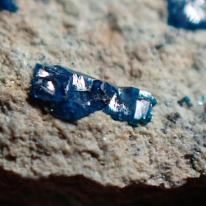 May include: Close-up of a vibrant blue mineral crystal fragment with reflective surfaces, set against a rough, light brown and gray rock matrix. The crystal's color is a deep, saturated blue.