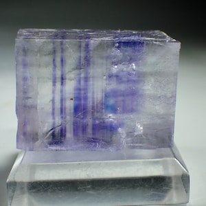 May include: A clear, translucent cube-shaped crystal with purple and blue striations. The crystal is sitting on a clear glass base.