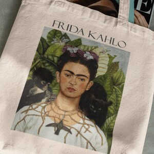 Könnte beinhalten: Beige Tragetasche mit einem Porträt von Frida Kahlo und dem Text "FRIDA KAHLO" darüber. Das Kunstwerk zeigt Kahlo mit einem Affen und einer schwarzen Katze. Die Tasche enthält Zeitschriften.