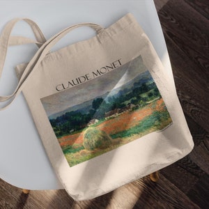 Può includere: Borsa tote in tela beige con un dipinto di paesaggio di Claude Monet. L'opera raffigura un campo con covoni, alberi e un cielo nuvoloso. Le parole "CLAUDE MONET" sono stampate in nero.