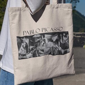 May include: A beige canvas tote bag featuring the name "PABLO PICASSO" above a black and white image of Picasso's artwork. The bag has long straps and is being carried outdoors.
