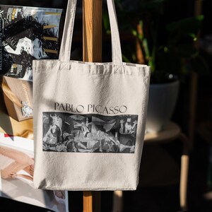 May include: Cream-colored tote bag featuring the name "PABLO PICASSO" in dark text above a black and white image of a painting. The bag has two handles and is hanging on a wooden support.