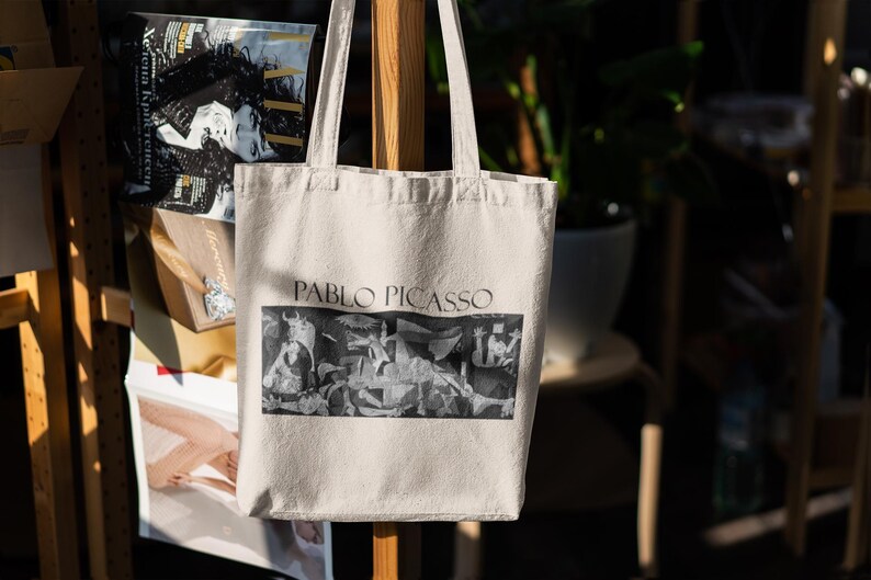 May include: Cream-colored tote bag featuring the name "PABLO PICASSO" in dark text above a black and white image of a painting. The bag has two handles and is hanging on a wooden support.