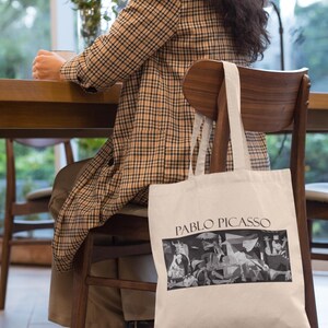 May include: A cream-colored tote bag featuring a black and white Pablo Picasso artwork. The bag has the text "PABLO PICASSO" printed in black. The bag is hanging on the back of a wooden chair.