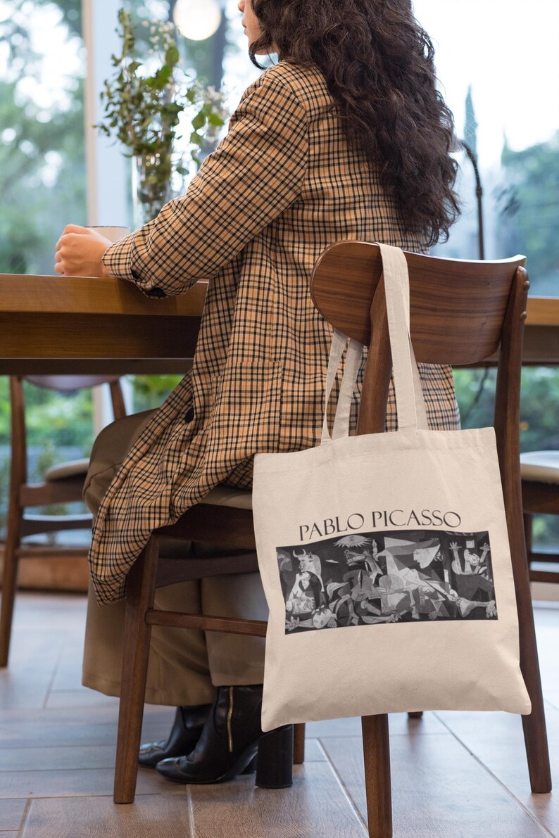 May include: A cream-colored tote bag featuring a black and white Pablo Picasso artwork. The bag has the text "PABLO PICASSO" printed in black. The bag is hanging on the back of a wooden chair.