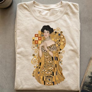 May include: A beige crew neck t-shirt featuring a central graphic print of Gustav Klimt's Portrait of Adele Bloch-Bauer I. The artwork displays a figure in a gold-toned, patterned gown with geometric shapes, swirls, and dark blue accents.