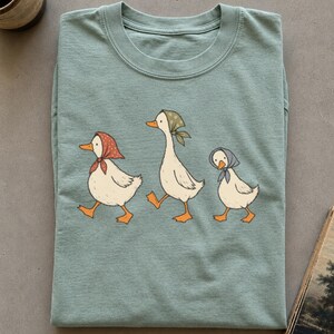 Puede incluir: Una camiseta de cuello redondo en color verde salvia con una ilustración gráfica de tres patos blancos caminando en fila. Cada pato lleva un pañuelo de diferente color en rojo, verde y azul, con picos y patas de color naranja brillante.