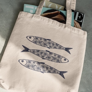 May include: A beige tote bag featuring three stylized fish illustrations. Each fish is decorated with a floral pattern in navy blue. The bag is filled with magazines and has a simple handle design.