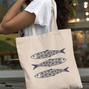 May include: A beige tote bag featuring three stylized fish. Each fish has a dark blue outline and is filled with a floral pattern. The bag is carried by a person wearing a white t-shirt.
