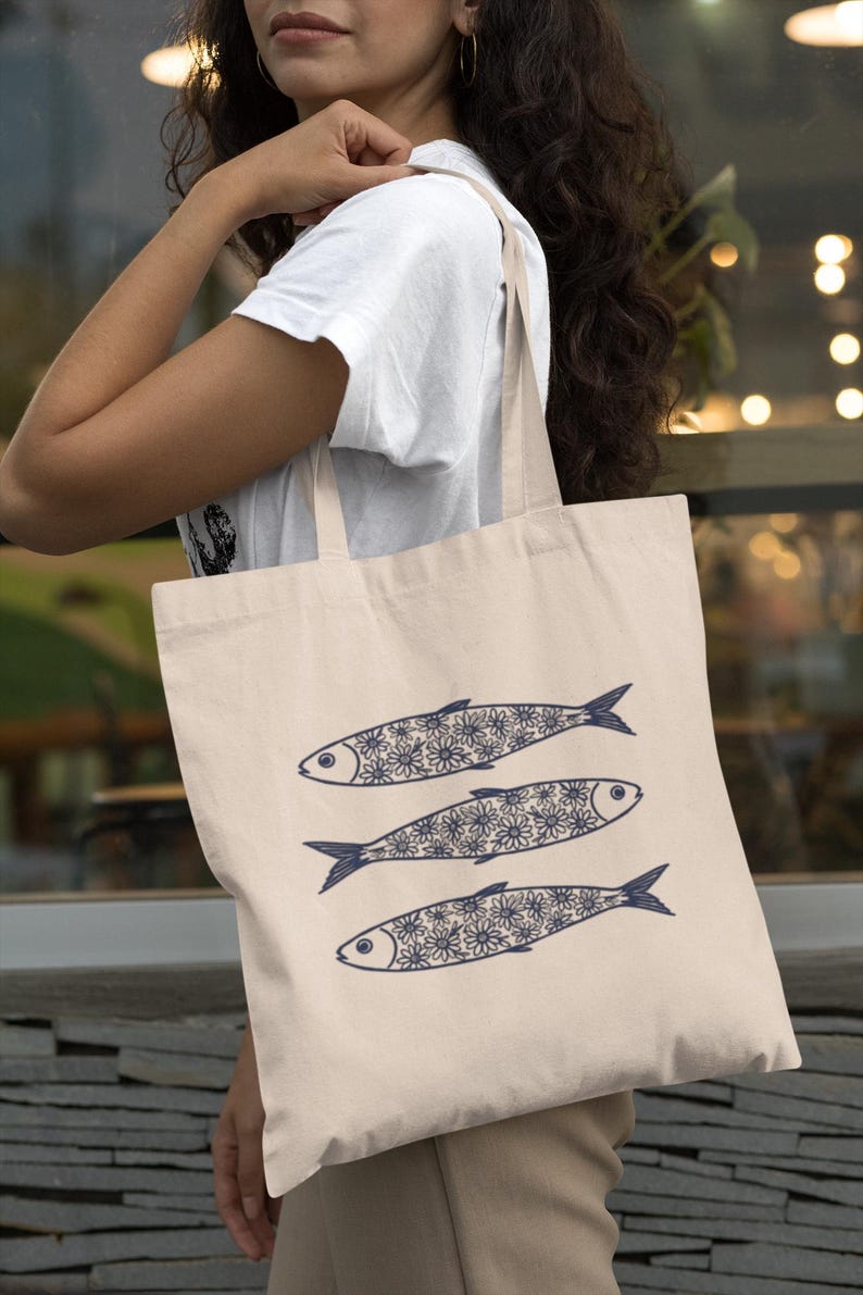 May include: A beige tote bag featuring three stylized fish. Each fish has a dark blue outline and is filled with a floral pattern. The bag is carried by a person wearing a white t-shirt.