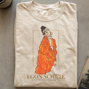 以下が含まれることがあります： ベージュのクルーネックTシャツ。エゴン・シーレの作品「Kneeling Female in Orange Red Dress, 1910」のグラフィックプリントが施されています。オレンジ色のローブをまとった人物のイラストが中央に描かれ、細い金色の長方形で囲まれています。