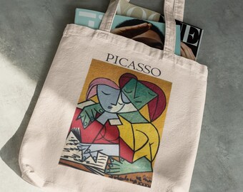 Puede incluir: Bolso tote beige con una impresión de una obra de arte de Picasso. La impresión representa dos figuras en un estilo cubista, con la palabra "PICASSO" encima de la imagen. La bolsa está llena de revistas.
