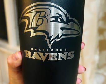 May include: Black stainless steel tumbler with a silver engraved Baltimore Ravens logo. The logo features a raven with the letter "B" on its chest and the words "Baltimore Ravens" underneath.