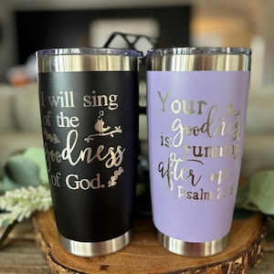May include: Two stainless steel tumblers with inspirational quotes. The black tumbler reads "I will sing of the goodness of God." The purple tumbler reads "Your goodness is running after me Psalm 23:6".