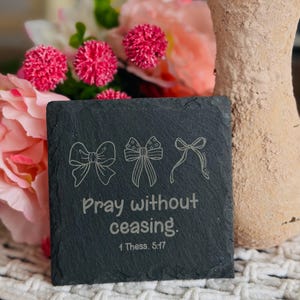 May include: A square, dark gray slate coaster with three engraved bow designs and the text "Pray without ceasing. 1 Thess. 5:17". The coaster is surrounded by pink flowers and a beige vase.
