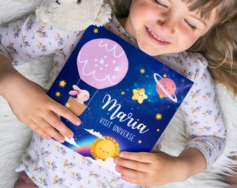 Puede incluir: Un libro azul con un globo aerostático rosa, estrellas y planetas. El libro dice "Maria Visit Universe" en la portada.