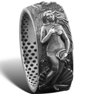Può includere: Anello color argento con un rilievo dettagliato di una figura femminile classica, forse Venere, che emerge da una conchiglia. L'anello ha una fascia strutturata con una serie di piccoli fori. Il design è intricato e artistico.