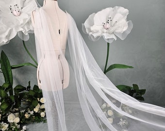 May include: A white tulle bridal cape draped over a dress form. The cape is sheer and has a delicate, flowing design. It is perfect for adding a touch of elegance to any wedding dress.