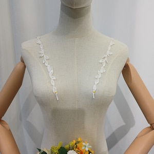 May include: A mannequin displays a white bridal bodice with delicate, leaf-shaped lace straps. The straps are adorned with small, sparkling embellishments and yellow accents. A colorful bouquet of flowers, including sunflowers and white blossoms, is held in front.