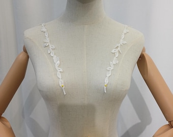 May include: A mannequin displays a white bridal bodice with delicate, leaf-shaped lace straps. The straps are adorned with small, sparkling embellishments and yellow accents. A colorful bouquet of flowers, including sunflowers and white blossoms, is held in front.