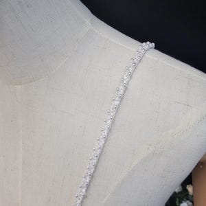 May include: Close-up of a white dress strap adorned with small, round, pearl-like beads. The strap is attached to a light-colored fabric, possibly a dress or top, displayed on a mannequin. The background is dark, highlighting the delicate details.
