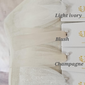 May include: Close-up of fabric swatches in light ivory, blush, champagne, and glitter light ivory. The swatches are attached to white cards with pearl pins. The fabric is draped over a mannequin.