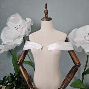 May include: White off-the-shoulder bridal bolero with a satin finish. The bolero is displayed on a white mannequin with wooden arms.