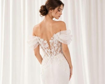 May include: A white off-the-shoulder gown featuring a sheer bodice with intricate floral lace embroidery and delicate tulle sleeves. The back of the dress showcases a deep V-shaped cutout, highlighting the detailed lace work and flowing fabric.