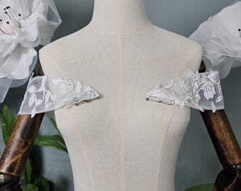 May include: A pair of white off-the-shoulder lace appliques with a floral design. The appliques are attached to a white dress form with safety pins.