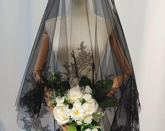 May include: A black tulle veil with intricate lace detailing, draped over a mannequin. The veil is paired with a bouquet of white roses and daisies, accented with green foliage. The lace design is floral.
