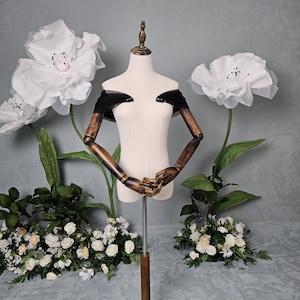 May include: A beige dress form with wooden arms and a wooden base. The dress form has a black off-the-shoulder design. Large white paper flowers and floral arrangements are in the background.